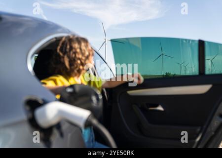 Eine Frau mit gebogenen Haaren, die ein gelbes Hemd trägt, sitzt in einem Elektroauto. Windräder im Hintergrund. Stockfoto