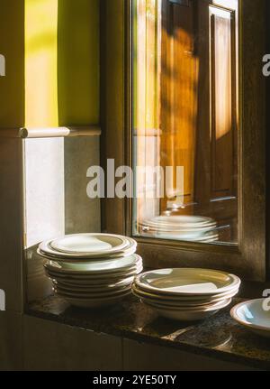 Stapel weißer Teller auf einer Granittheke, in warmes Sonnenlicht getaucht, das durch ein Holzfenster filtert. Stockfoto