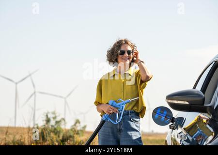 Die Frau hält eine Wasserstoff-Tankdüse. Betanken des Autos mit Wasserstoffkraftstoff. Windräder im Hintergrund. Stockfoto