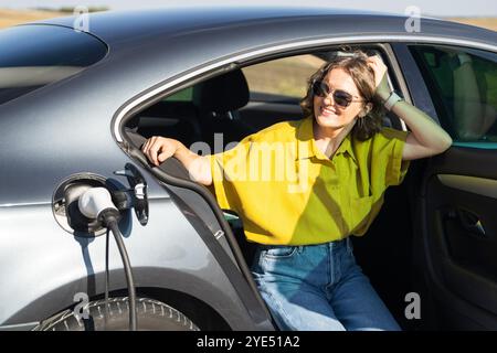 Eine Frau mit gebogenen Haaren, die ein gelbes Hemd trägt, sitzt in einem Elektroauto. Windräder im Hintergrund. Stockfoto