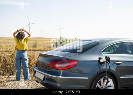 Frau mit gebogenen Haaren, die ein gelbes Hemd trägt, steht neben dem Aufladen des Elektroautos. Windräder im Hintergrund. Stockfoto