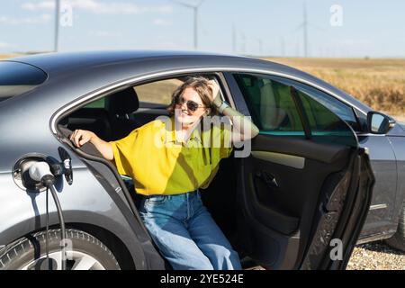 Eine Frau mit gebogenen Haaren, die ein gelbes Hemd trägt, sitzt in einem Elektroauto. Windräder im Hintergrund. Stockfoto