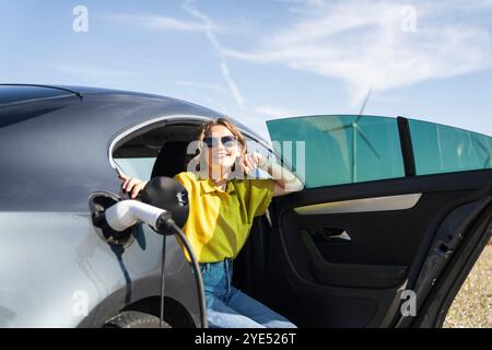 Eine Frau mit gebogenen Haaren, die ein gelbes Hemd trägt, sitzt in einem Elektroauto. Windräder im Hintergrund. Stockfoto