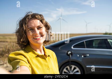 Frau mit gebogenen Haaren, die ein gelbes Hemd neben dem Aufladen des Elektroautos trägt. Windräder im Hintergrund. Stockfoto