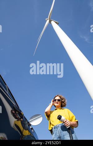 Eine Frau mit gewölbten Haaren, die ein gelbes Hemd und eine Sonnenbrille trägt, steht neben dem Aufladen des Elektroautos. Windkraftanlage im Hintergrund. Stockfoto
