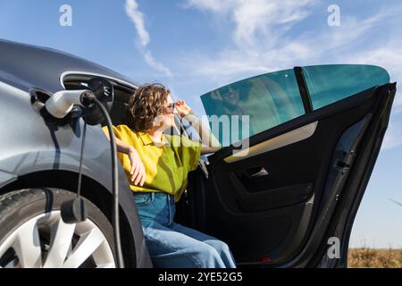 Eine Frau mit gebogenen Haaren, die ein gelbes Hemd trägt, sitzt in einem Elektroauto. Windräder im Hintergrund. Stockfoto