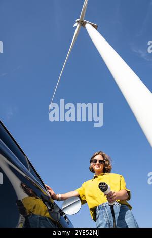 Eine Frau mit gewölbten Haaren, die ein gelbes Hemd und eine Sonnenbrille trägt, steht neben dem Aufladen des Elektroautos. Windkraftanlage im Hintergrund. Stockfoto