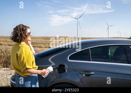 Eine Frau mit gewölbten Haaren, die ein gelbes Hemd und eine Sonnenbrille trägt, steht neben dem Aufladen des Elektroautos. Windkraftanlage im Hintergrund. Stockfoto