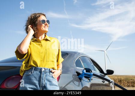 Die Frau hält eine Wasserstoff-Tankdüse. Betanken des Autos mit Wasserstoffkraftstoff. Windräder im Hintergrund. Stockfoto