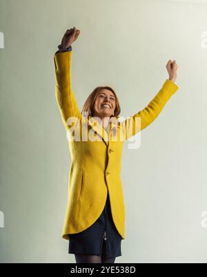 Eine begeisterte Frau in einem gelben Mantel hebt ihre Arme zur Feier und drückt Freude und Begeisterung über einen Erfolg oder eine Leistung aus Stockfoto