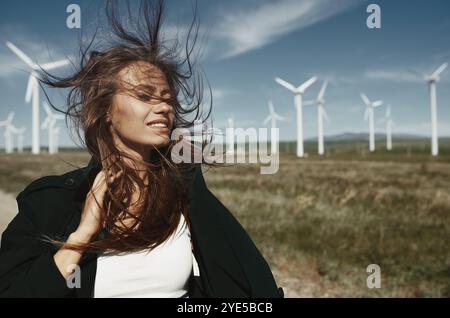 Eine fröhliche junge Frau, im Wind flatterndes Haar, lächelt strahlend vor dem Hintergrund hoch aufragender Windturbinen auf einem weiten, offenen Feld Stockfoto