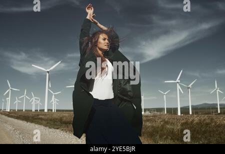 Eine junge Frau, die in einem modernen dunklen Mantel gekleidet ist, tanzt anmutig vor einer rauen Landschaft, die mit aufragenden Windturbinen unter einem dramatischen bewölkten Himmel gesäumt ist Stockfoto