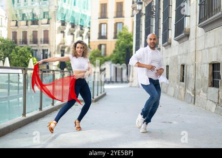 Ein energiegeladenes Paar spielt lateinischen Tanz auf einer sonnigen Straße. Die Frau wirbelt einen roten Schal, während beide fröhliche Ausdrücke zeigen, die den vib umarmen Stockfoto