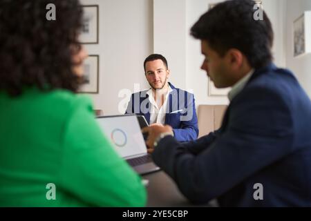 Drei Männer sitzen an einem Tisch mit Laptop und Handy. Ein Mann schaut auf den Laptop, während die anderen zwei ihn anschauen. Die Szene ist Profis Stockfoto