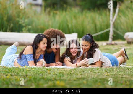 Vier junge Frauen liegen auf einer Decke im Park, eine hält ein Handy. Sie scheinen die Gesellschaft des anderen zu genießen und eine Pause einzulegen Stockfoto