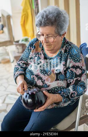 Eine ältere Frau, die Aubergine schält, sitzt auf der Terrasse ihres Gartens in Sizilien. Stockfoto