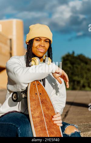 Eine lächelnde junge schwarze Lateinfrau trägt eine gelbe Mütze und ein lässiges Outfit, hält ein Skateboard und posiert draußen mit Kopfhörern Stockfoto