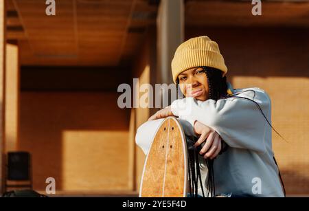 Ein fröhliches junges Mädchen mit Vitiligo, geflochtenen Haaren und einer hellgelben Mütze sitzt auf ihrem Skateboard auf einem sonnigen Campus, ihr angenehmer Ausdruck r Stockfoto