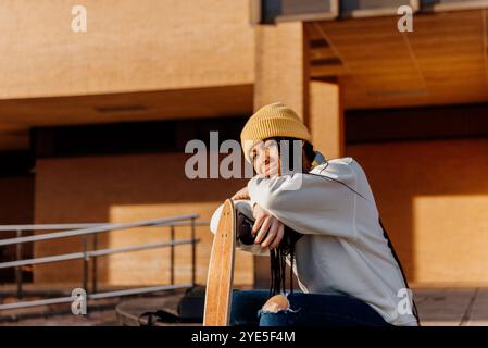 Eine fröhliche junge Lateinfrau mit Vitiligo, eine gelbe Mütze und lässige Kleidung, sitzt mit ihrem Skateboard auf einem sonnigen Campus, embraci Stockfoto