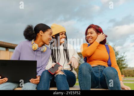 Drei lateinische Frauen sitzen draußen auf einem Campus, führen lebhafte Gespräche und Lachen, mit Laptops und Smartphones und präsentieren einen Moment Stockfoto