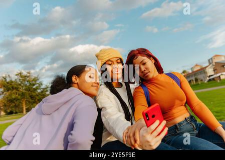 Drei lebendige junge Frauen, die ein Smartphone verwenden und sich auf einem sonnigen Campus-Rasen in zwangloser Atmosphäre unterhalten, stellen Jugend und Verbindung in einem lebendigen Ambiente dar Stockfoto