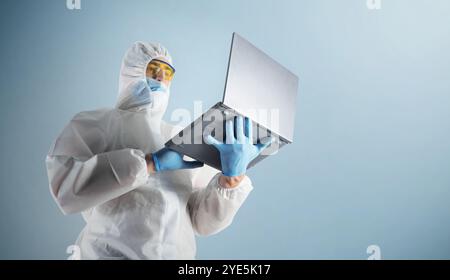 Laborarbeiter, ein Mediziner in Schutzanzug, Handschuhen und Schutzbrille, benutzt einen Laptop auf blauem Hintergrund Stockfoto