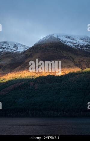 Goldenes Sonnenlicht bricht über den zerklüfteten schottischen Highlands auf und beleuchtet die schneebedeckten Gipfel und dichten Wälder darunter Stockfoto