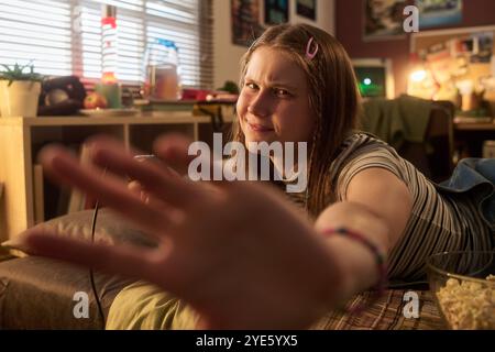 Junge Frau, die in einem gemütlichen Zimmer sitzt, mit einem unordentlichen Schreibtisch im Hintergrund, die Hand zur Kamera streckt und einen verspielten Ausdruck gibt. Zimmer mit warmer Beleuchtung und persönlichen Retro-Gegenständen Stockfoto