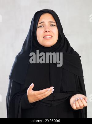 Die junge muslimische Frau in schwarzem Hijab ist über schlechte Nachrichten verärgert und hält ihre Hände dejektiv hoch. Stockfoto
