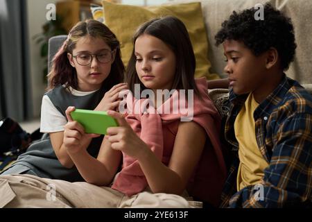 Gruppe von drei Kindern aus verschiedenen Hintergründen, dicht auf der Couch sitzend und konzentriert auf den Smartphone-Bildschirm. Ein Kind hält ein hellgrünes Smartphone Stockfoto