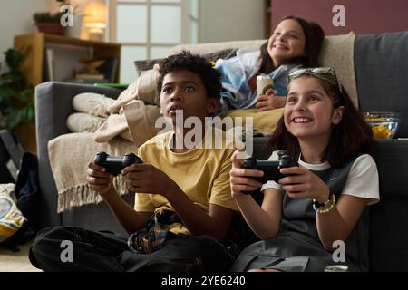 Drei Kinder spielen Videospiele auf der Couch, während sie lachen und Spaß haben. Regale mit Büchern und Dekorationen im Hintergrund schaffen eine gemütliche Atmosphäre Stockfoto