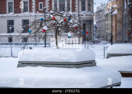 Ein kleiner Baum, der mit roten und blauen Ornamenten verziert ist, hebt sich vor dem Hintergrund von schneebedeckten Bänken und Gebäuden hervor, während Schneeflocken sanft fallen Stockfoto