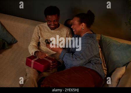 Hochwinkel-Porträt eines glücklichen afroamerikanischen erwachsenen Paares, das Geschenke an Weihnachten austauscht und eine gemütliche Feier zu Hause mit schwachem Licht im Kopierraum genießt Stockfoto