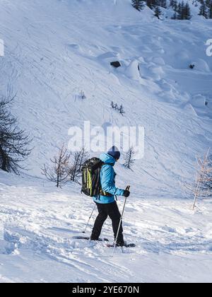 Ein Wanderer in einer blauen Jacke mit Rucksack und Trekkingstöcken führt über einen verschneiten Berghang. Schneebedeckte Bäume und zerklüftetes Gelände sorgen für eine Winterwerbung Stockfoto