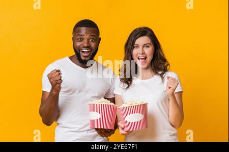 Fröhliches interracial Paar jubelt mit Popcorn-Eimern und weckt Fäuste in Aufregung Stockfoto