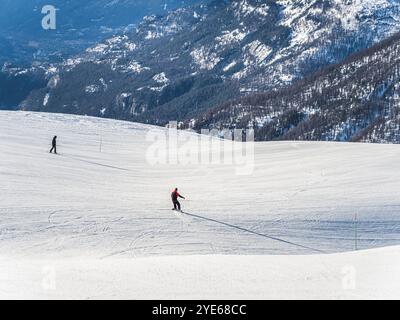 Zwei Skifahrer gleiten einen sanften, schneebedeckten Berghang hinunter, umgeben von malerischem alpinen Gelände. Das Bild erfasst den offenen Raum, den scharfen Schnee und V Stockfoto