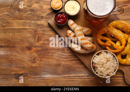 Leckere bayerische Würstchen, Saucen, Sauerkraut, Brezeln und Becher Bier auf hölzernem Hintergrund. Oktoberfest-Feier Stockfoto