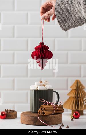 Heißer Kakao im festlichen Motto mit Marshmallows in einem Becher und einer Frauenhand, die ein Weihnachtsbaumspielzeug hält. Stockfoto