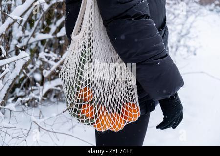 Unerkennbare Frau, die reife Mandarinen in wiederverwendbarem Netzbeutel hält, draußen im verschneiten Winterwald. Konzept nachhaltiger Weihnachtsfeier, Verantwortung und Inklusivität. Orangen im Baumwollbeutel Stockfoto