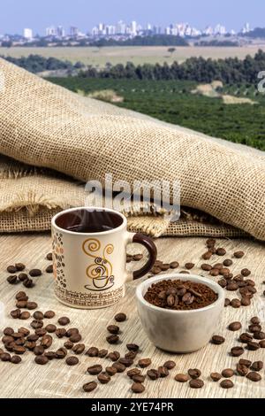 Eine Tasse frischen Kaffee und geröstete Bohnen in einer Tüte und einem Topf auf dem Tisch vor dem Hintergrund einer Kaffeeplantage und Skyline-Stadt in Brasilien Stockfoto