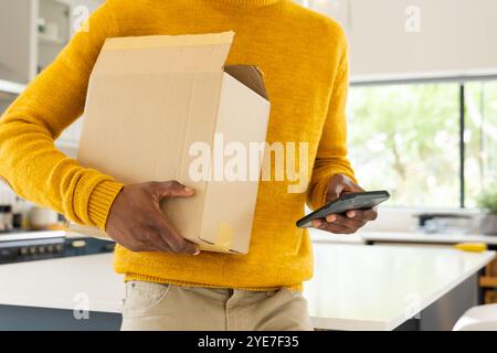 afroamerikanischer Mann, der Karton hält und das Smartphone zu Hause nutzt, um effizient multitasken zu können Stockfoto