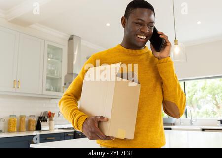 Lächelnder afroamerikaner, der Karton hält, während er in der Küche mit dem Smartphone spricht, zu Hause Stockfoto