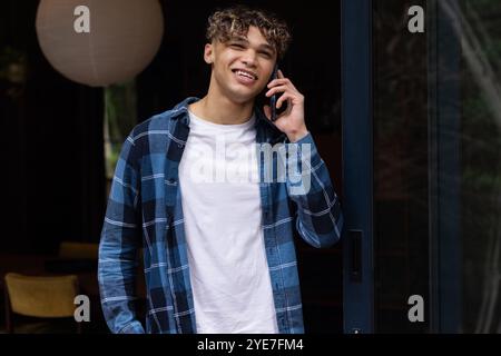 Junger Mann in kariertem Hemd, der auf dem Smartphone spricht und zu Hause lächelt Stockfoto