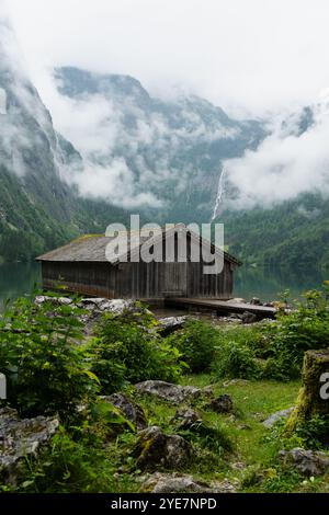 Rustikale Holzhütte am Ufer des Obersees, Nationalpark Berchtesgaden, Deutschland, umgeben von nebeligen Bergen und üppigem Grün. Stockfoto