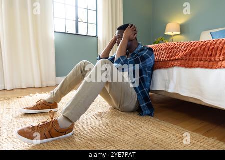 afroamerikaner sitzt auf dem Schlafzimmerboden, hält den Kopf in den Händen, fühlt sich gestresst, zu Hause Stockfoto