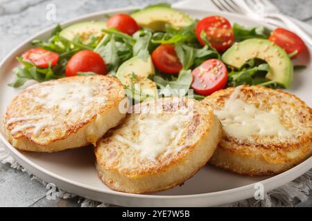 Cheesy Eggy Crumpets mit Salat aus Avocado, Tomaten, Rucola und Spinat in Nahaufnahme auf dem Teller auf dem Tisch. Horizontal Stockfoto