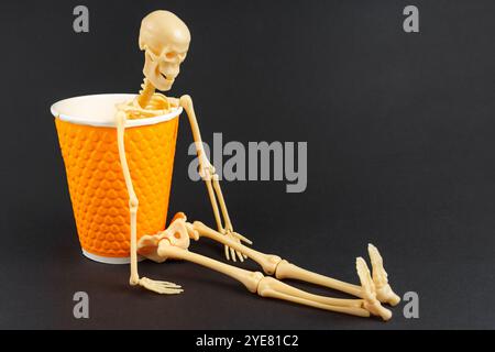 Gebrochenes Skelett in oranger Tasse vor schwarzem Hintergrund, symbolisiert Erschöpfung und Bedarf an Kaffee, um Energie wiederzubeleben. Stockfoto