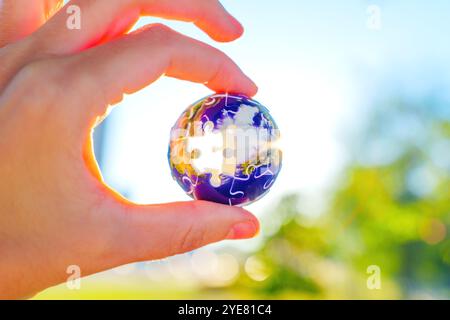 Hand hält ein kugelförmiges 3D-Puzzle mit verschwommenem natürlichen Hintergrund, das globale Einheit und Problemlösung symbolisiert. Stockfoto