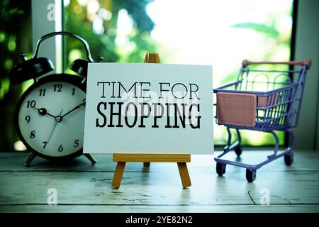 Time for Shopping geschrieben auf Papierkarte auf Holzstaffelei mit Wecker und Mini-Einkaufswagen im Fensterhintergrund Stockfoto