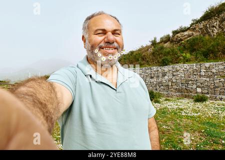 Lachender reifer Mann mit Gänseblümchen im Bart, der Selfie macht. Stockfoto